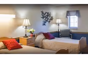 A cozy bedroom in a senior living facility with two single beds, each with beige bedding and red pillows. There are two bedside tables with lamps and a small flower arrangement on one table. A decorative metal wall art piece is mounted above the beds, and a window with a blue valance allows natural light into the room.