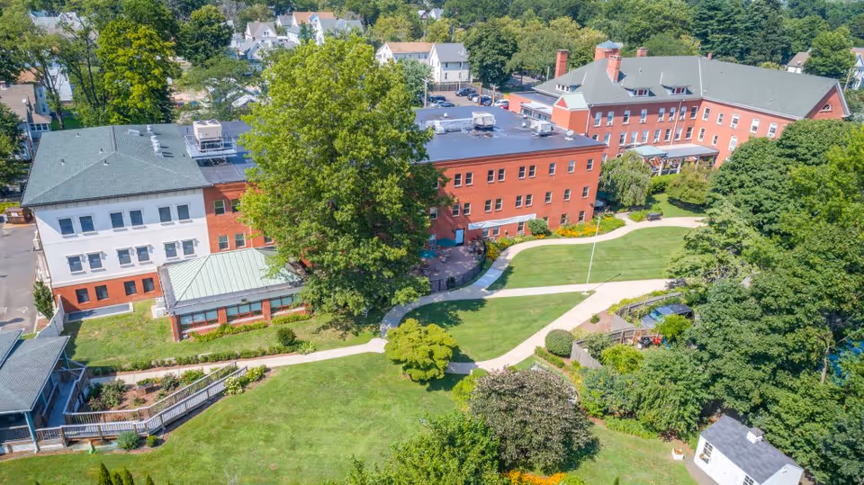 Aerial view of a red-brick senior living complex with green lawns, winding walkways, trees, and surrounding houses.