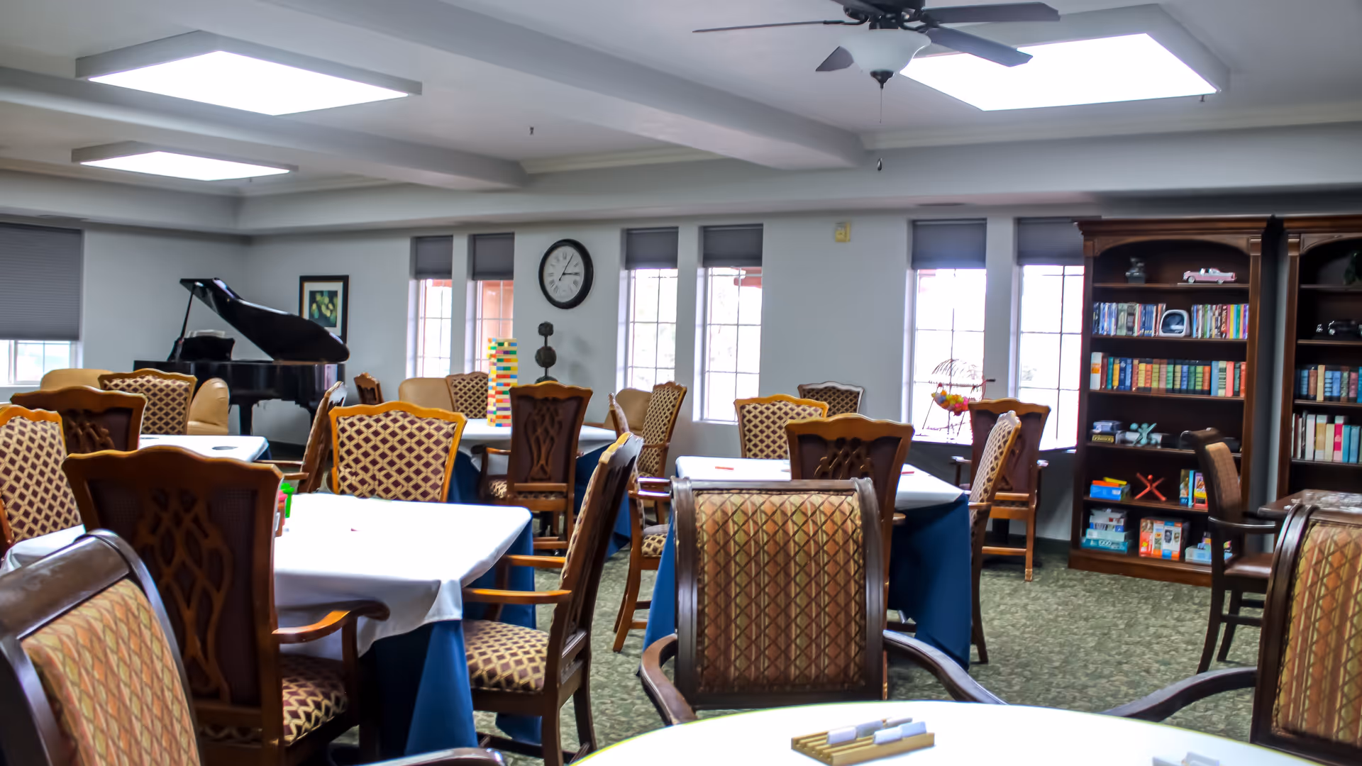 A bright senior living common room with multiple tables covered in white and blue tablecloths surrounded by patterned chairs. A grand piano is visible in the back left corner. There are several windows with blinds, a clock on the wall, and a bookshelf filled with books and board games on the right side.