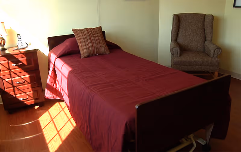 Sunlit bedroom with a single bed covered in a burgundy bedspread, a wooden nightstand, and an upholstered armchair.