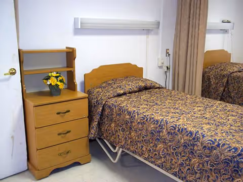 A small bedroom with a single bed covered in a patterned bedspread. Next to the bed is a wooden nightstand with three drawers and a small shelf on top holding a pot of yellow flowers. A beige curtain hangs beside the bed, and a mirror reflects part of the room.