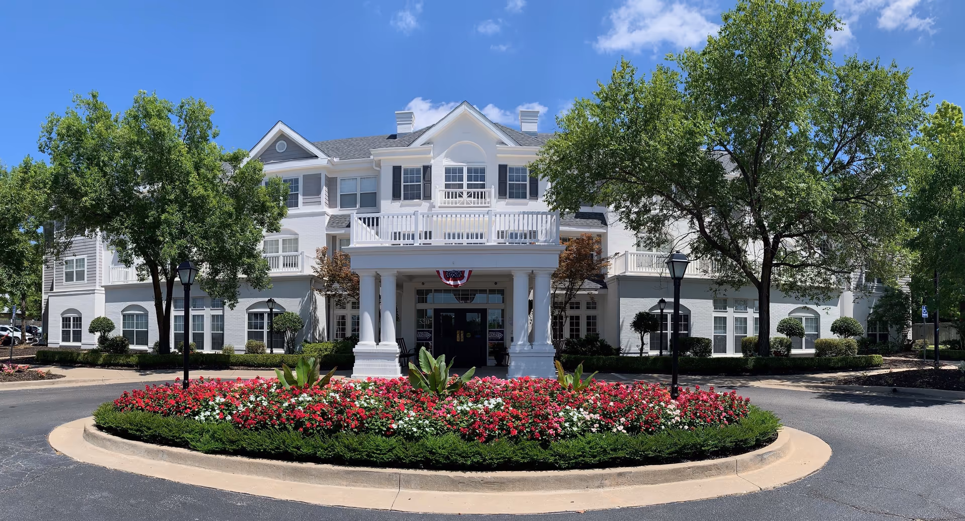 Front exterior view of The Grove at Midtown, a large white multi-story building with black shutters, surrounded by green trees and a circular flower bed with red and white flowers in front of the entrance.