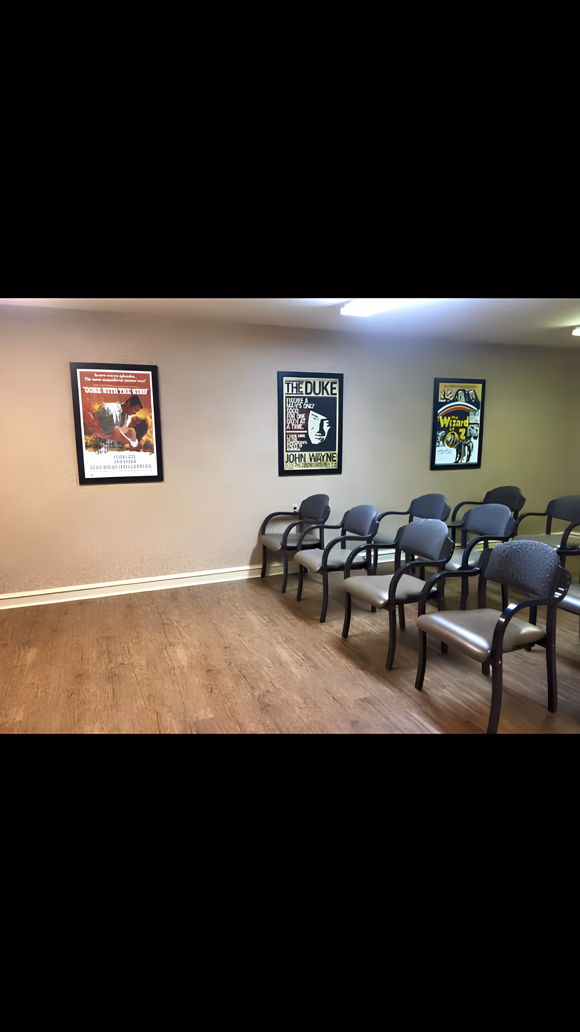 A room with several rows of gray chairs with armrests arranged facing the left side. The walls are beige and decorated with three framed vintage-style movie posters. The floor has a wood-like finish and the room is lit by ceiling lights.