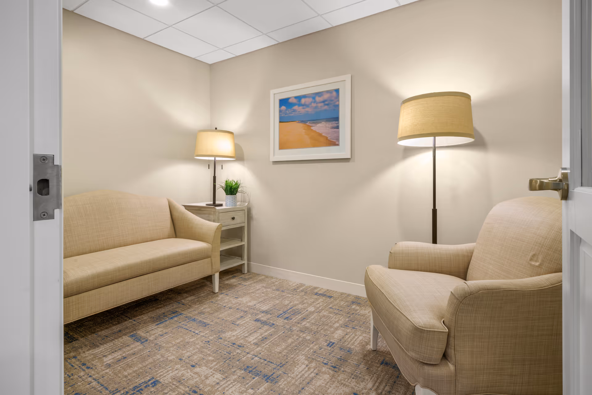 Small neutral-toned sitting room with a beige sofa and armchair, side table, two lamps, and a framed beach picture on the wall.