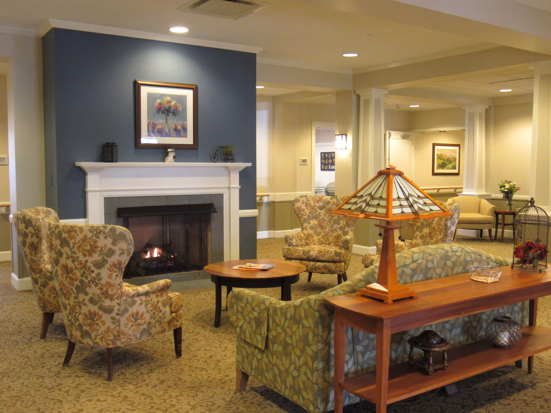 A cozy living room area in a senior living facility with a lit fireplace set against a blue accent wall. The room features floral patterned armchairs, a green leaf-patterned sofa, a wooden coffee table with magazines, and a wooden console table with a decorative lamp and ornaments. The walls are light-colored with framed artwork, and the space is softly lit with recessed ceiling lights.