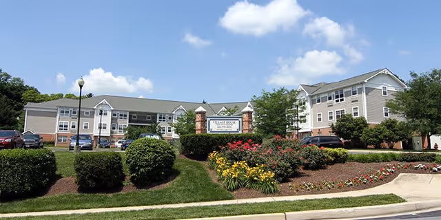 Exterior view of Village House Apartments, a multi-story residential building with beige siding and brick accents, surrounded by landscaped bushes, flowers, and a parking area with several cars under a blue sky with scattered clouds.