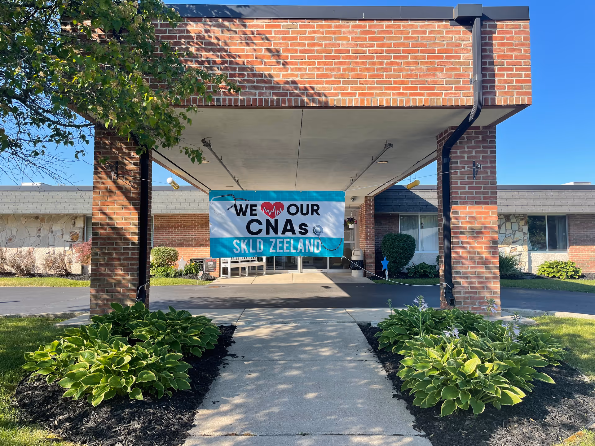 Entrance of a brick building with a covered driveway. A banner hanging under the canopy reads 'We love our CNAs SKLD Zeeland' with a heart and stethoscope graphic. There are green plants and bushes on either side of the walkway leading to the entrance.