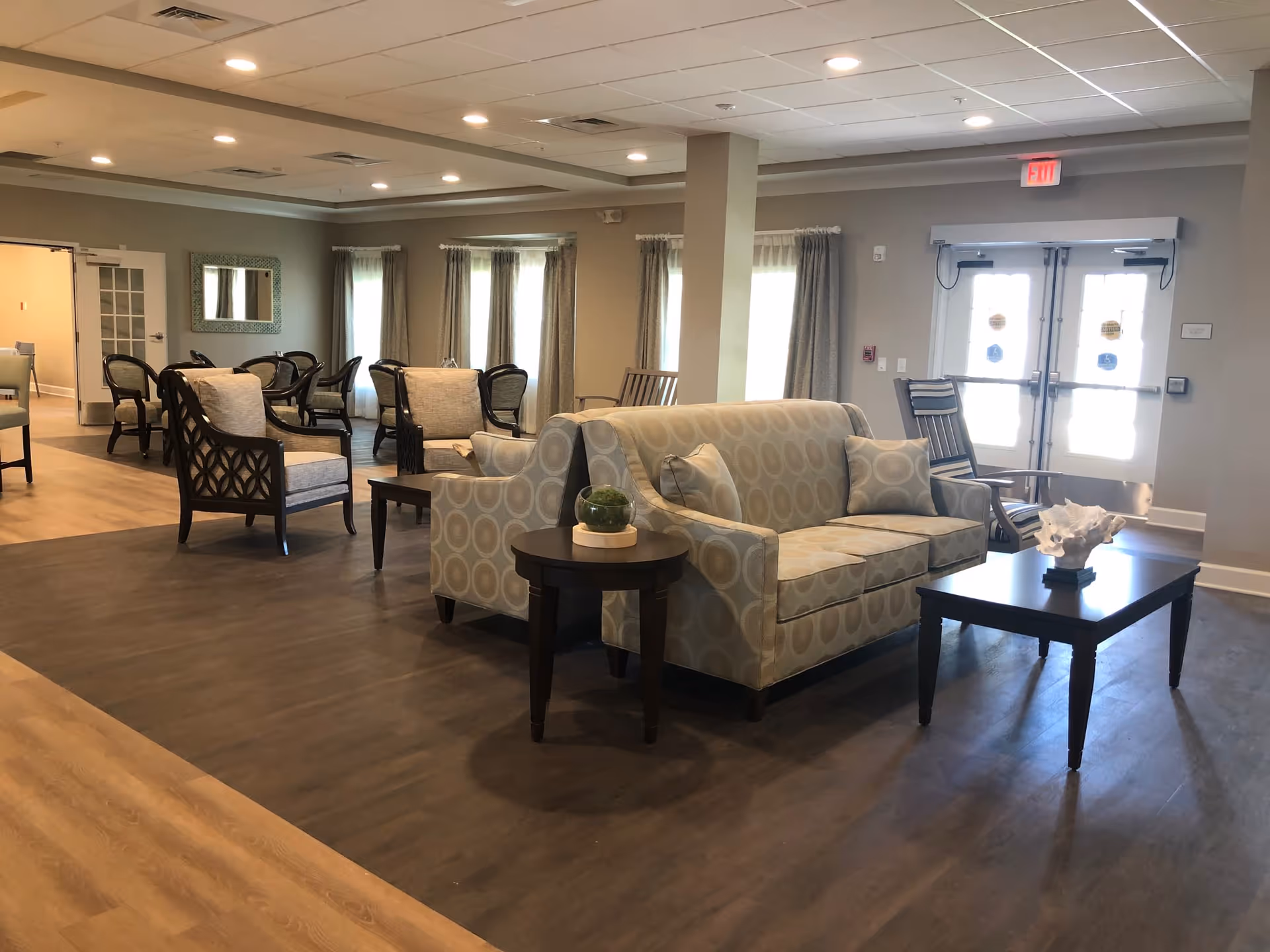 A spacious, well-lit common area with patterned sofas, armchairs, and wooden tables. The room has large windows with curtains, a double glass door exit, and a mix of wood and dark flooring. The ceiling has recessed lighting and a drop ceiling design.