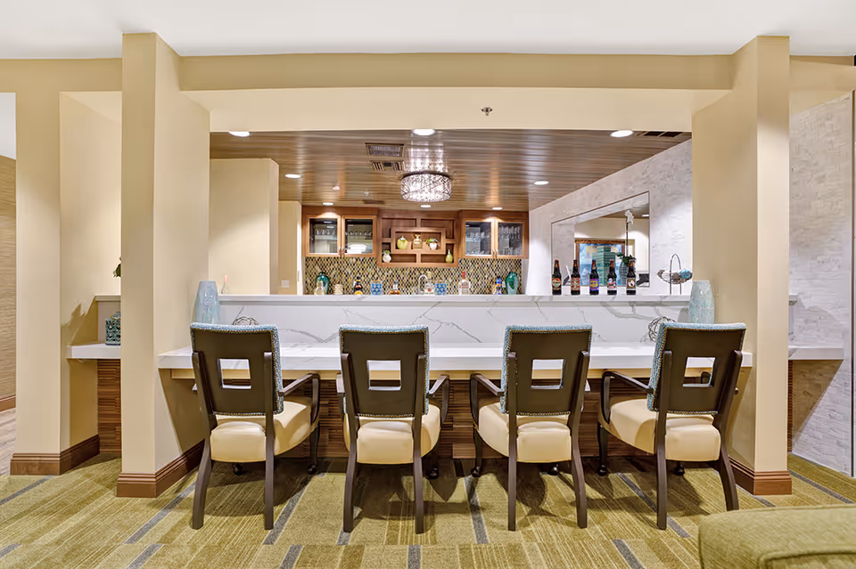 Interior view of a modern bar area with four cushioned chairs lined up at a white marble countertop. Behind the bar, there are wooden cabinets with glass doors, decorative vases, and bottles on display. The ceiling above the bar is wooden with recessed lighting and a decorative chandelier. The floor is carpeted in a green pattern, and the walls are painted in a light beige color.