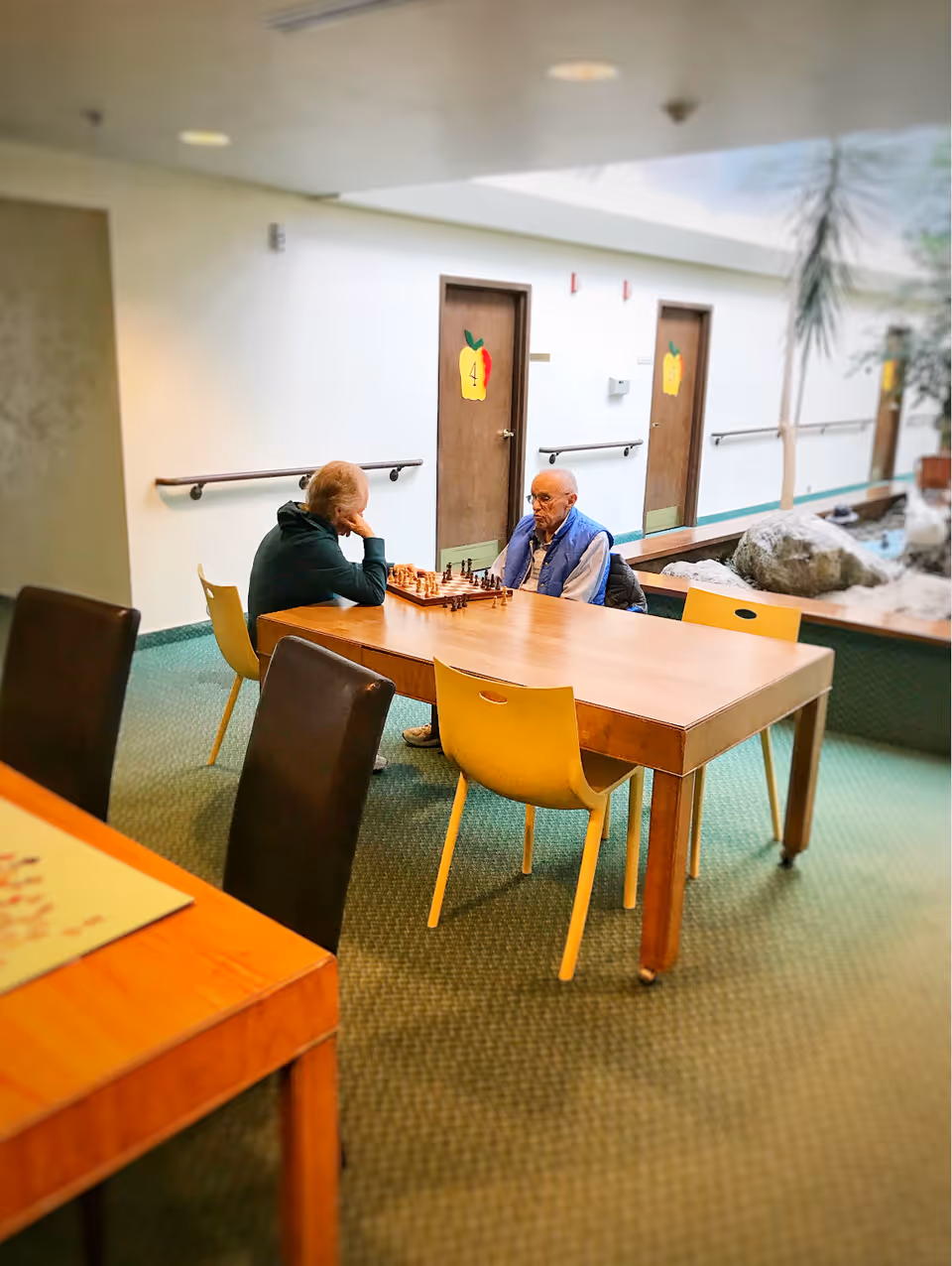 Two elderly men sitting at a wooden table playing chess in a well-lit indoor common area with green carpet. The background shows doors with colorful apple-shaped signs and a small indoor garden with rocks and plants.