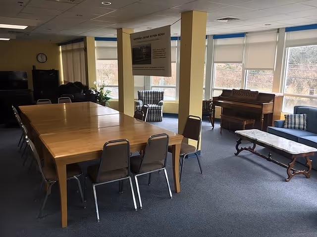 Spacious common room with a long wooden table and chairs, lounge seating, and a piano by large windows.