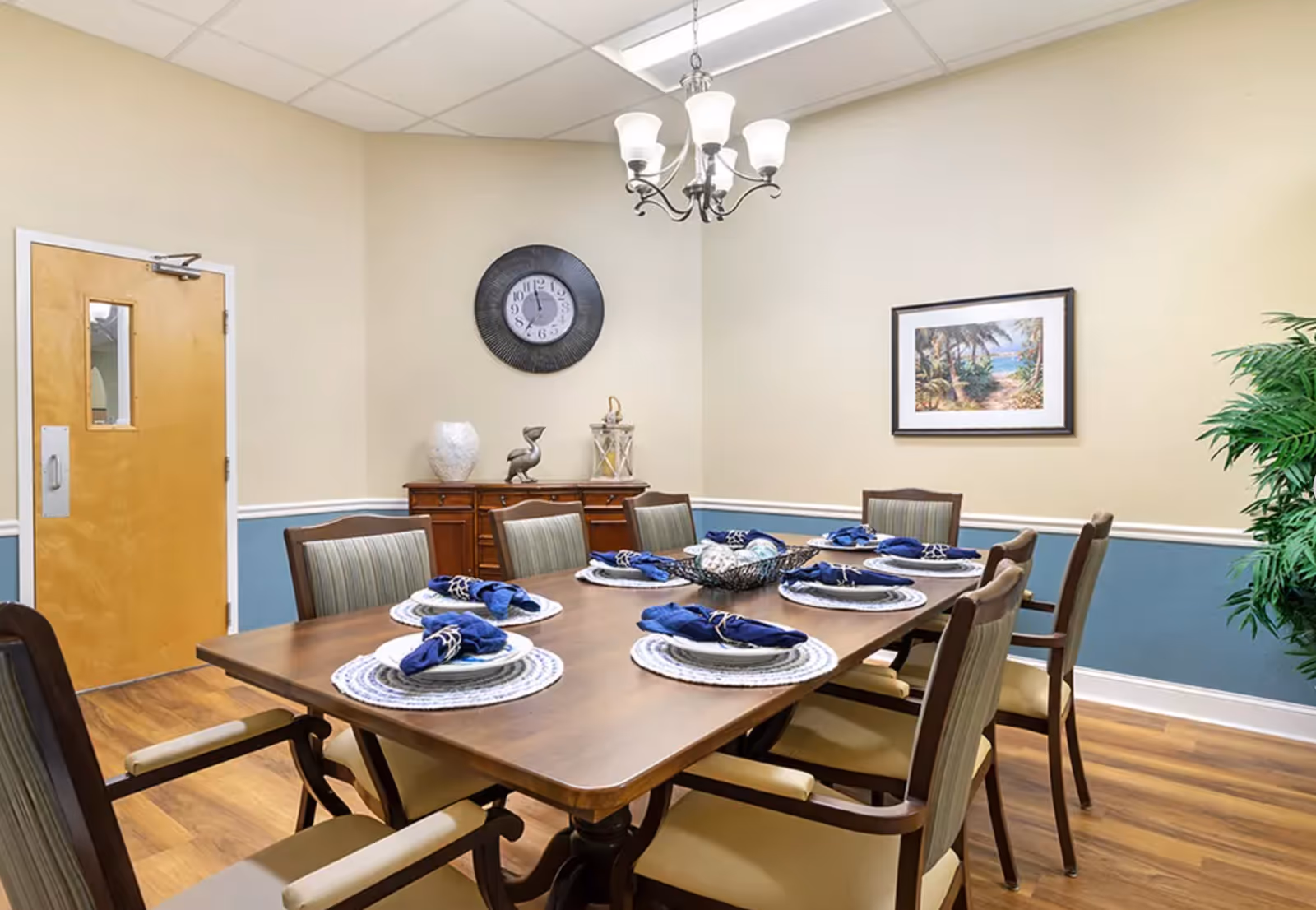 Well-appointed dining room with a long wooden table set for eight, blue napkins, a chandelier, wall clock, and framed artwork.