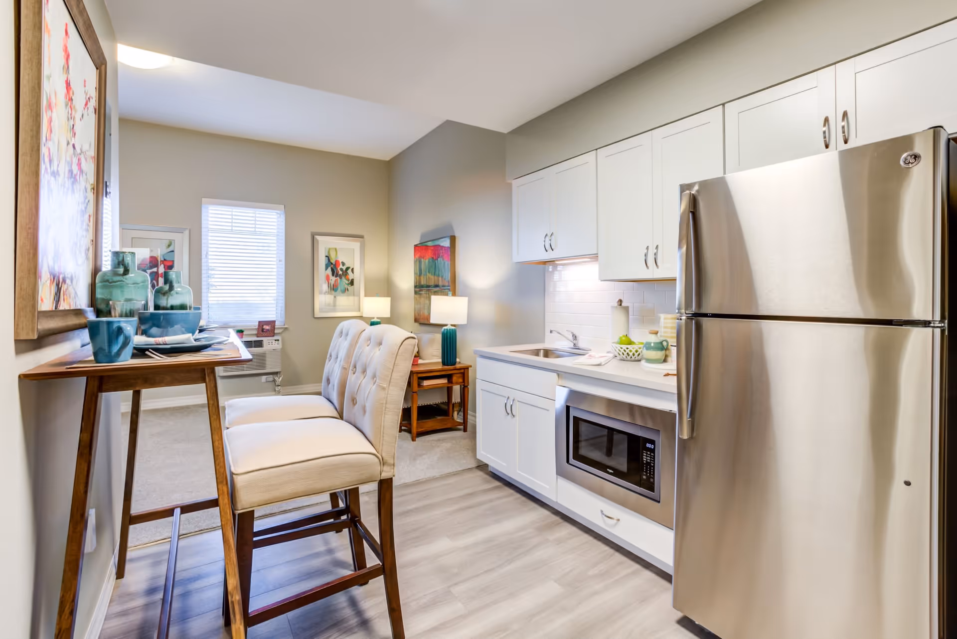 A modern kitchen area with white cabinets, a stainless steel refrigerator, a built-in microwave, and a sink. There is a small wooden dining table with two cushioned chairs. The kitchen opens into a living space with a side table, lamp, and framed artwork on the walls.