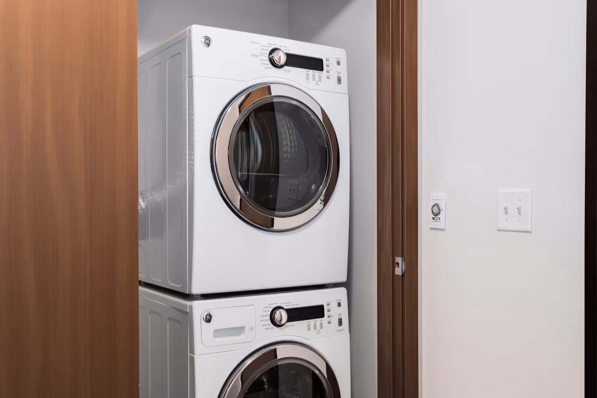 Stacked white front-loading washer and dryer units in a small laundry closet with wooden trim and white walls.