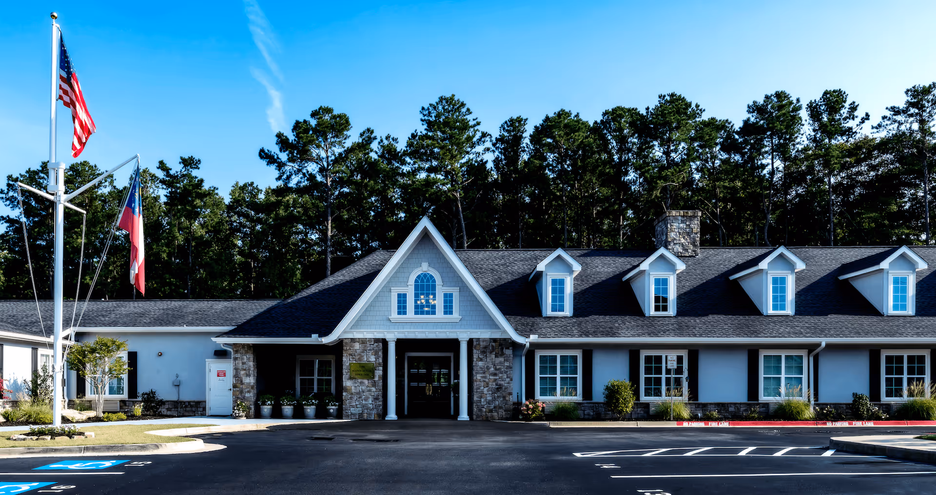 Front exterior view of The Mann House Cumming building with a stone and light blue facade, multiple windows, and a peaked roof. There are two flagpoles with the American flag and another flag, a parking lot with marked spaces including handicapped spots, and tall trees in the background under a clear blue sky.