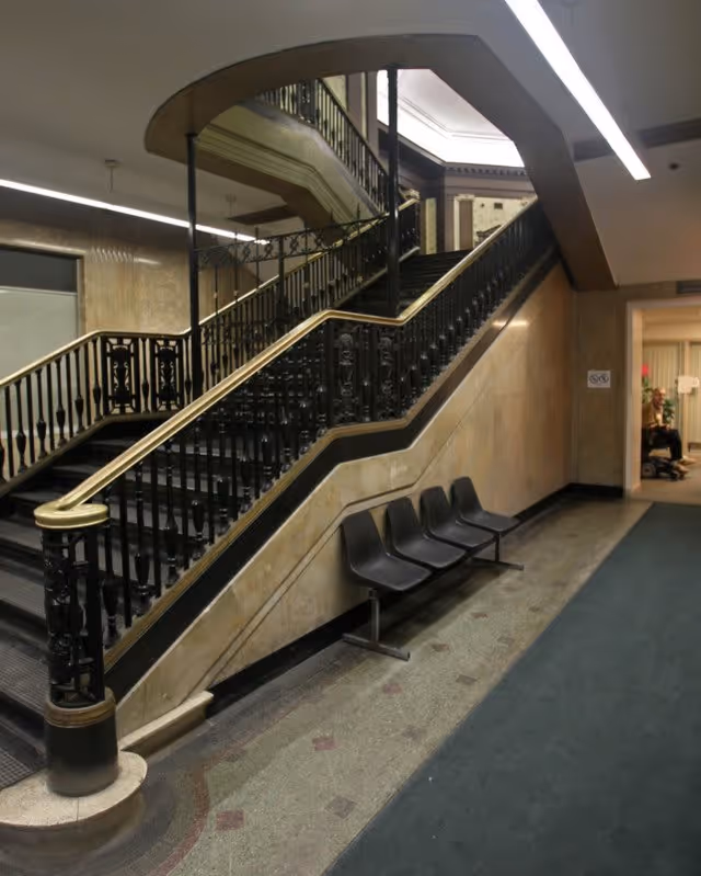 Interior view of a building hallway featuring a wide staircase with ornate black railings and a brass handrail. Four black plastic chairs are lined up against the wall beneath the staircase. The floor has a patterned tile design and a dark carpet runner. The walls are covered with beige marble or stone panels, and fluorescent lighting is visible on the ceiling.