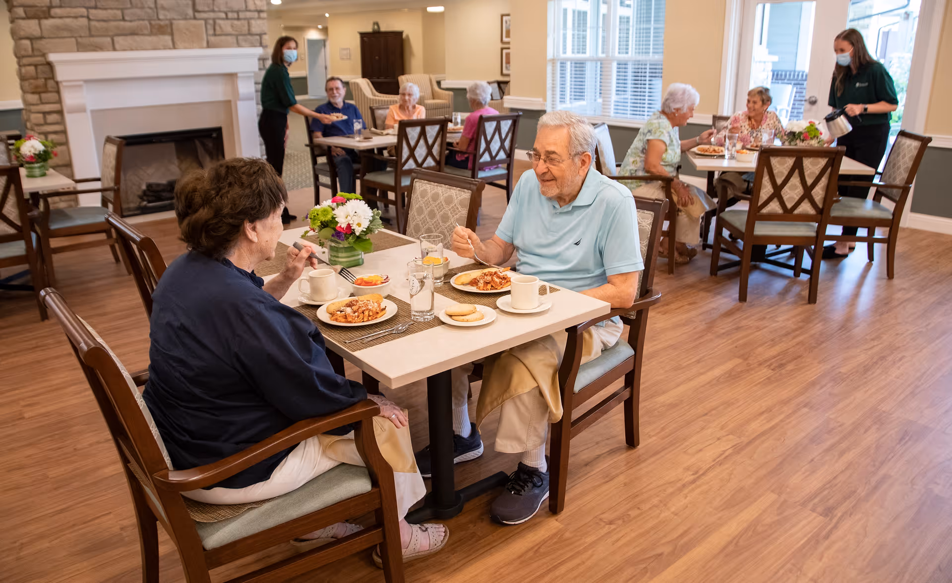 Several elderly people seated at tables in a dining room of a senior living facility, eating meals and engaging in conversation. Two staff members wearing masks are serving and assisting the residents. The room has wooden flooring, a fireplace, and large windows letting in natural light.