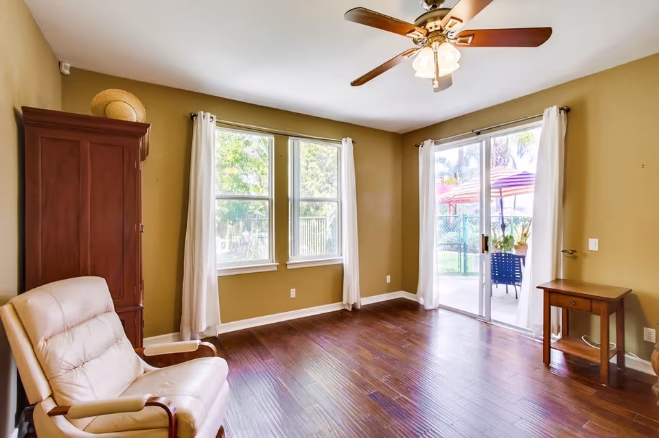 A bright room with wooden flooring, beige walls, and a ceiling fan with lights. There is a cream-colored cushioned armchair on the left, a tall wooden cabinet in the corner, and a small wooden side table on the right. Two windows with white curtains and a sliding glass door with white curtains lead to an outdoor area with patio furniture and umbrellas.