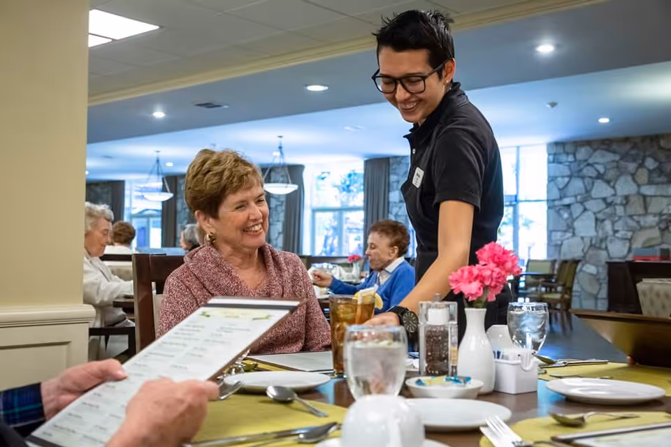 A smiling waitress serving a drink to a seated elderly woman in a dining room. Other elderly people are seated at tables in the background, and the room has large windows and a stone wall.