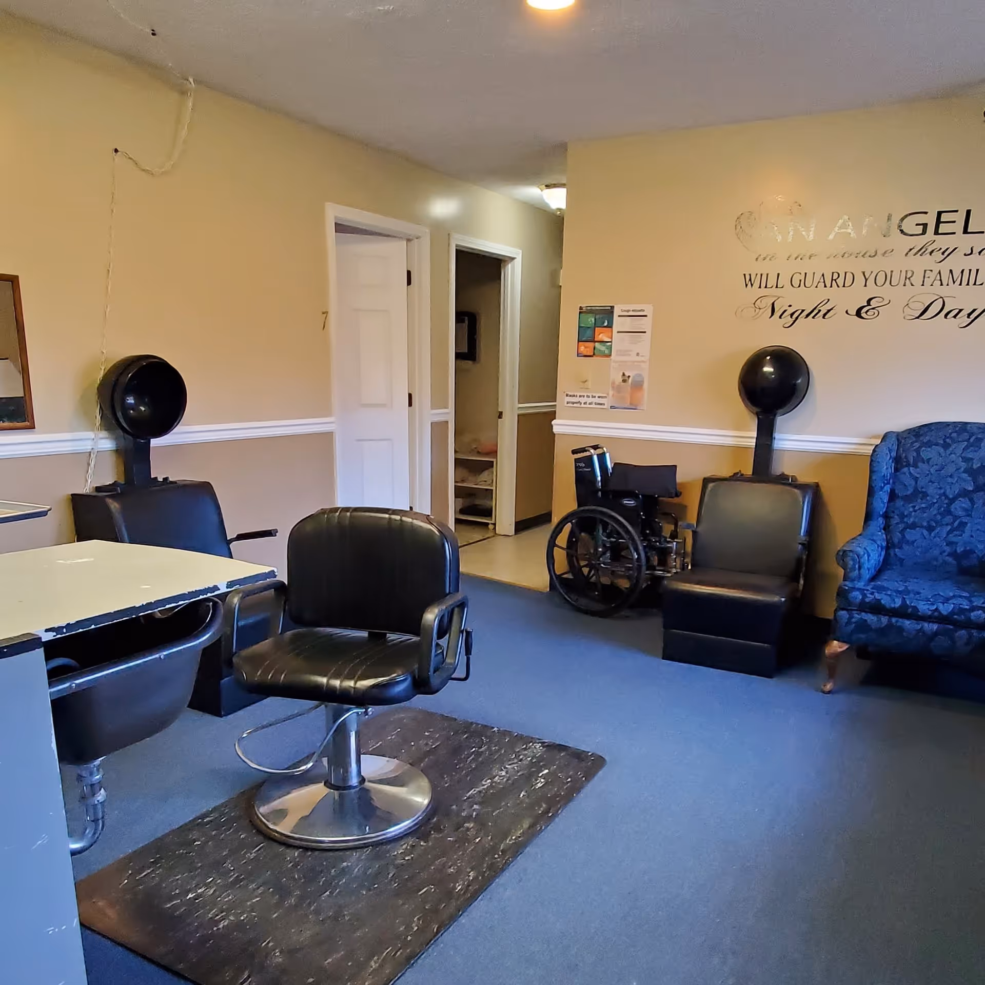 Interior room with salon chairs and hair dryers, a wheelchair, a blue upholstered armchair, and a wall with a partial quote about Angel Acres guarding families night and day.
