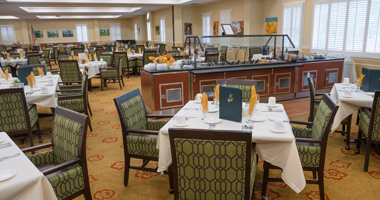 A spacious dining room with multiple tables covered in white tablecloths, each set with cups, plates, silverware, and yellow folded napkins. The chairs have green patterned upholstery. In the center is a buffet station with food trays and a glass sneeze guard. The room has large windows with white shutters and several paintings on the walls.
