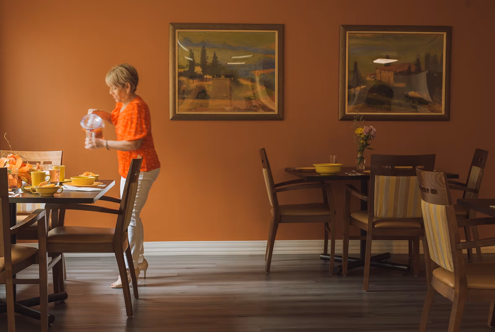 A woman pours water at a set dining table in a warm orange-walled dining room with wooden chairs and framed paintings.