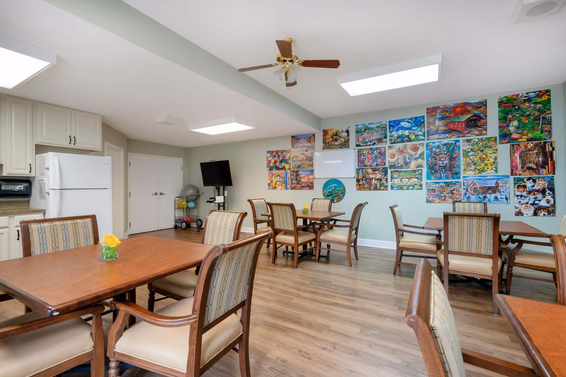 Bright communal dining and activity room with multiple wooden tables and chairs, a kitchenette with refrigerator, TV, and a wall covered in colorful artwork.