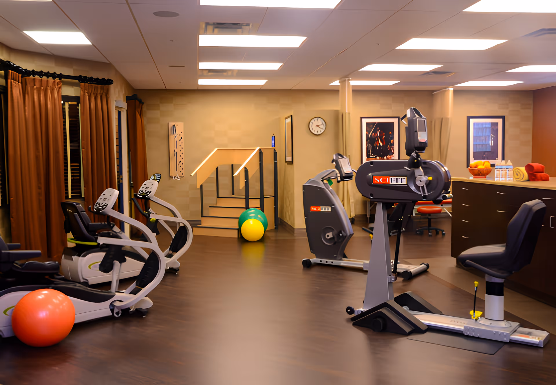 Well-lit indoor fitness room with cardio machines, exercise balls, and seating in a senior care facility.