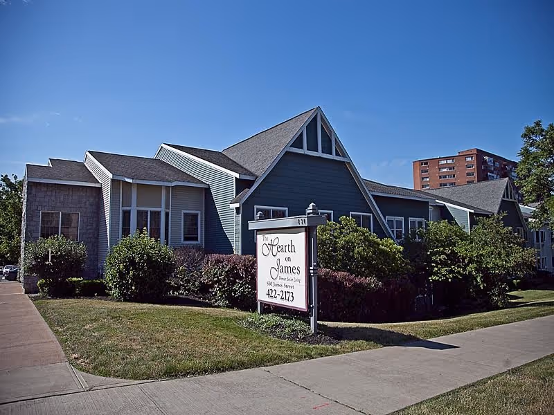 Exterior view of The Hearth on James senior living facility building with a sign in front displaying the facility name and contact number. The building has a blue-gray facade with white trim, multiple peaked roofs, and is surrounded by bushes and a well-maintained lawn under a clear blue sky.