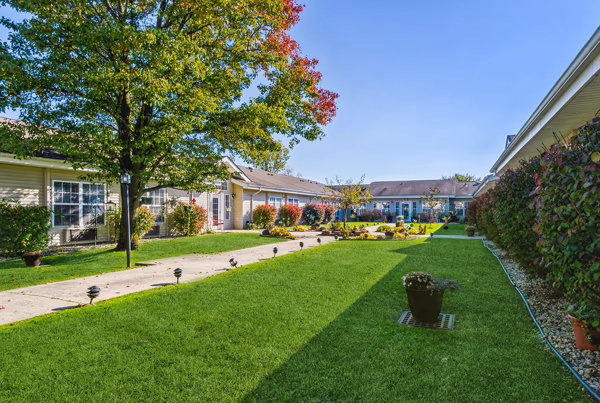 A sunny outdoor courtyard area with green grass, a paved walkway lined with small lights, and a variety of plants and trees. Surrounding the courtyard are single-story buildings with beige siding and multiple windows. The sky is clear and blue.