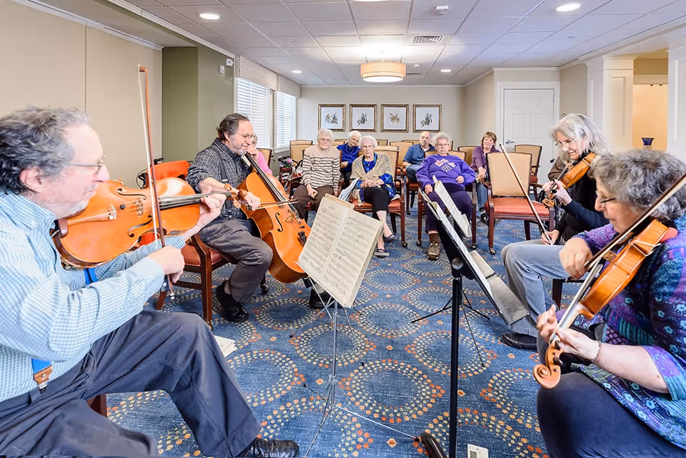 A group of elderly people seated in a room watching and listening to four musicians playing string instruments including violins and a cello. The room has patterned blue carpet, beige walls, and framed artwork on the back wall.