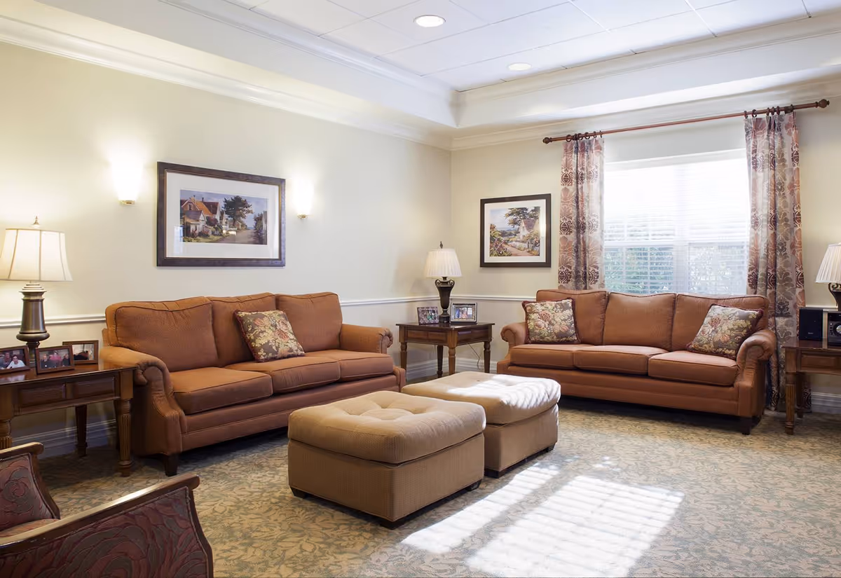 A bright, well-lit living room with two matching sofas, ottomans, side tables with lamps, framed artwork, and a window with curtains.