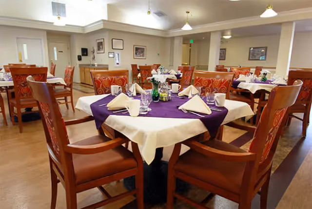 A dining room with multiple tables set for a meal. Each table is covered with a white tablecloth and a purple runner, and is set with folded napkins, glasses, mugs, and small condiment containers. The room has wooden chairs with red patterned cushions and a light-colored floor. The walls are decorated with framed pictures and the ceiling has recessed lighting and hanging fixtures.