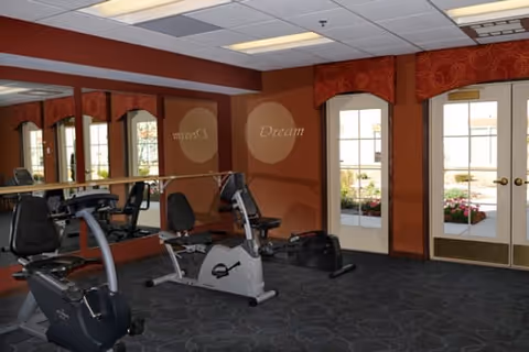Interior view of a fitness room with exercise bikes, large mirrors on one wall, and double glass doors leading outside. The room has a patterned carpet, warm-colored walls, and ceiling lights.