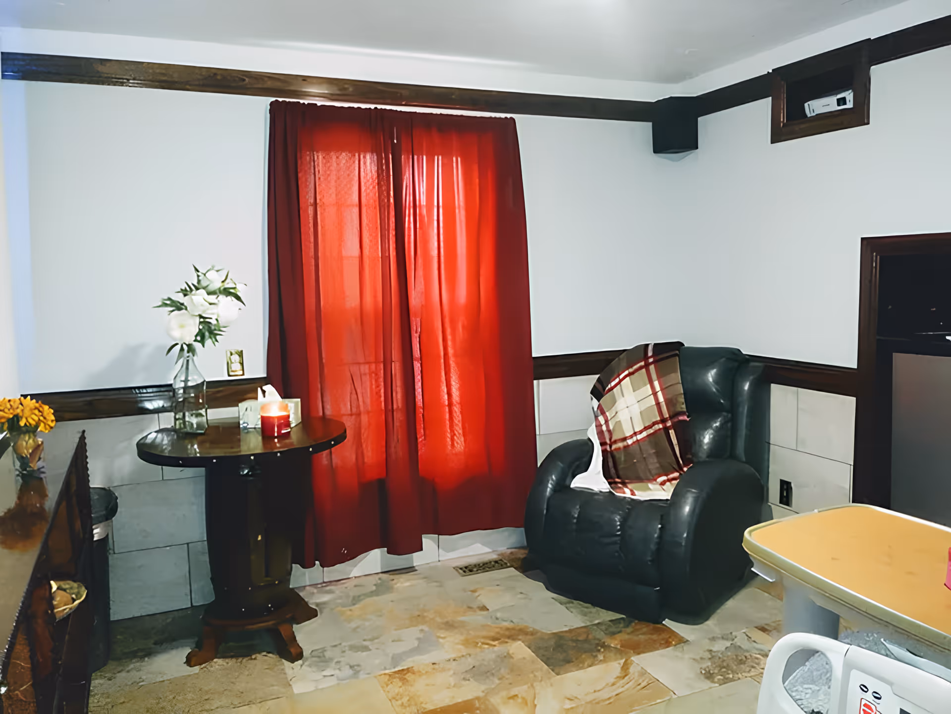 A cozy interior room with a dark green leather armchair draped with a plaid blanket, a small round wooden table with a vase of white flowers and a lit candle, red curtains covering a window, and a tiled floor.