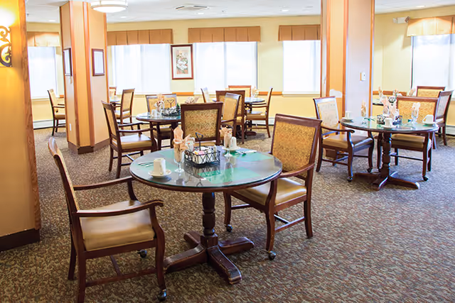 A bright dining room with multiple round tables set with cups, napkins, and utensils. The room has carpeted floors, large windows with beige valances, and wooden chairs with cushioned seats and backs. The walls are painted light yellow and there are decorative wall sconces and framed artwork.