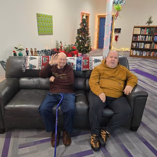 Two elderly men sitting on a dark gray leather couch in a decorated common area. One man is wearing a brown sweater and blue pants, holding a blue cane and waving, while the other man is wearing a mustard-colored hoodie and black pants. Behind them is a Christmas tree with lights and ornaments, holiday decorations on a table, a bookshelf filled with books, and a purple and gray patterned carpet.