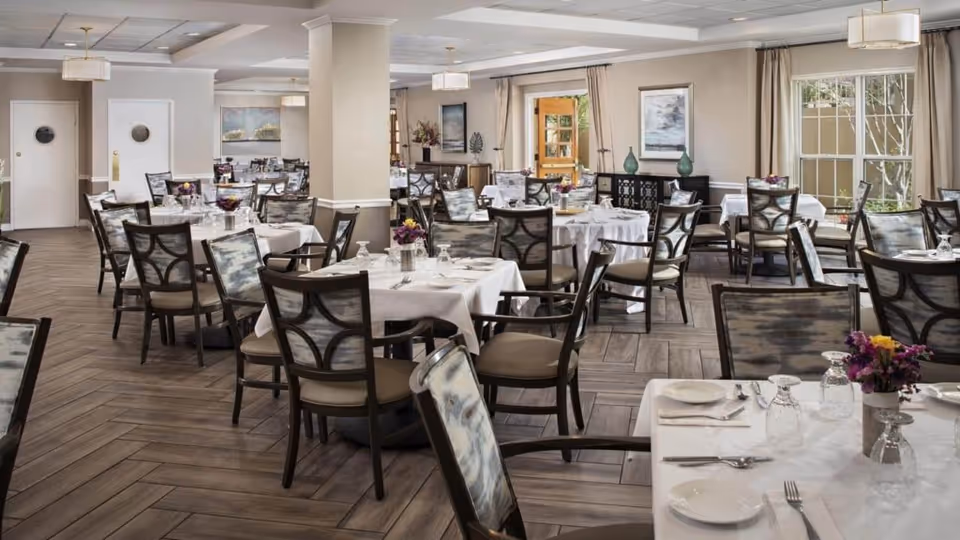 A spacious and elegant dining room with multiple tables covered in white tablecloths, each set with plates, glasses, and silverware. The chairs have patterned upholstery and wooden frames. The room features large windows with beige curtains, light fixtures hanging from the ceiling, and decorative artwork on the walls. The floor has a wood-like herringbone pattern.