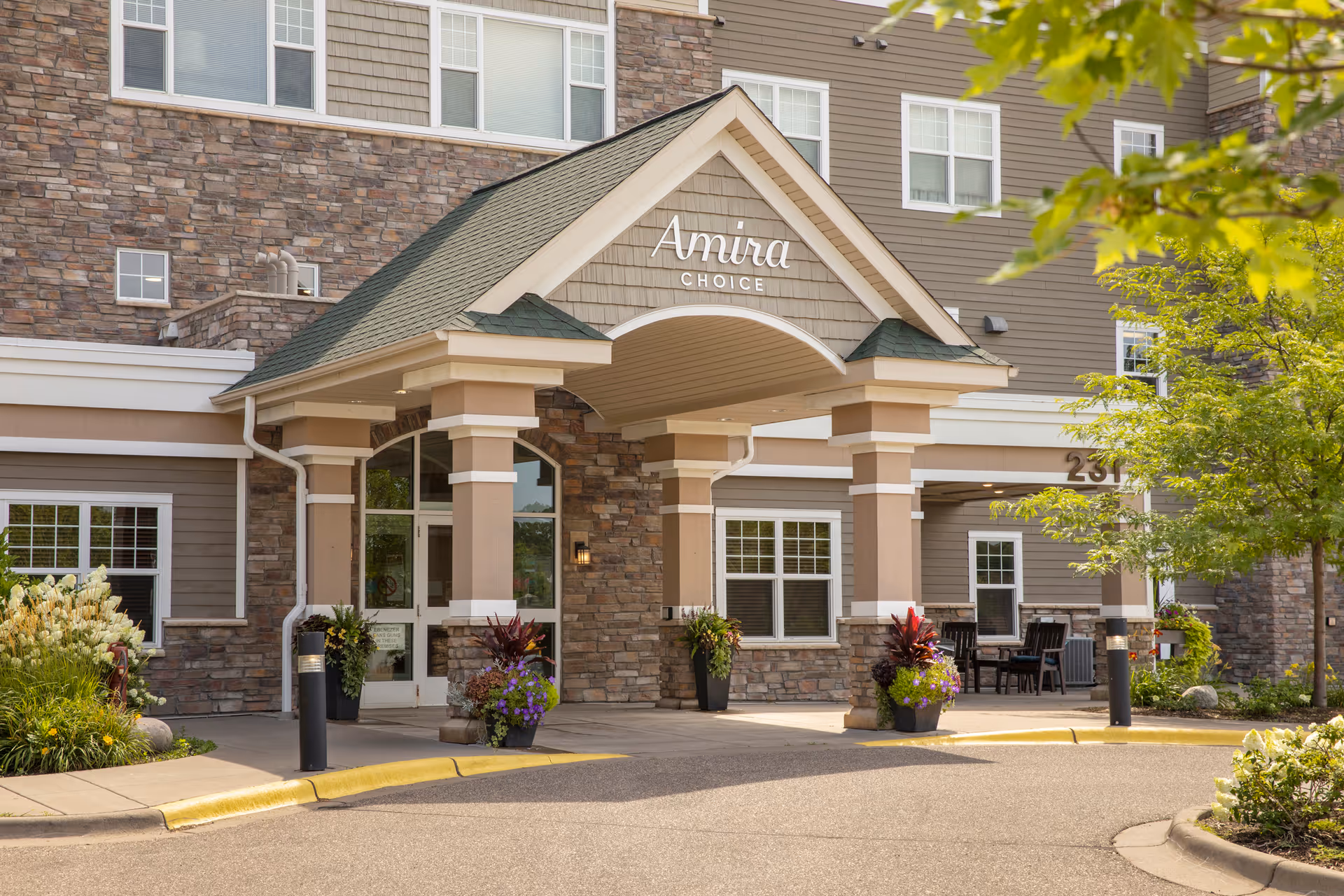 Entrance of Amira Choice Forest Lake facility with a covered driveway supported by columns, stone and siding exterior walls, potted plants, and a tree with green leaves on the right side.