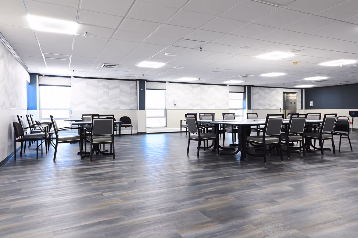 A spacious, well-lit room with multiple round and rectangular tables surrounded by chairs. The room has large windows with blinds, light-colored walls with a subtle pattern, and a wood-look floor. There is an elevator door visible on the far wall.