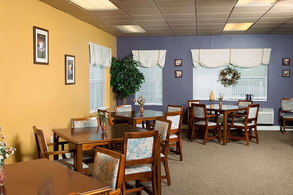A dining room with several wooden tables and chairs with floral upholstery. The walls are painted yellow and purple, decorated with framed pictures and a wreath. Windows have white blinds and valances, and there are small flower arrangements on the tables.