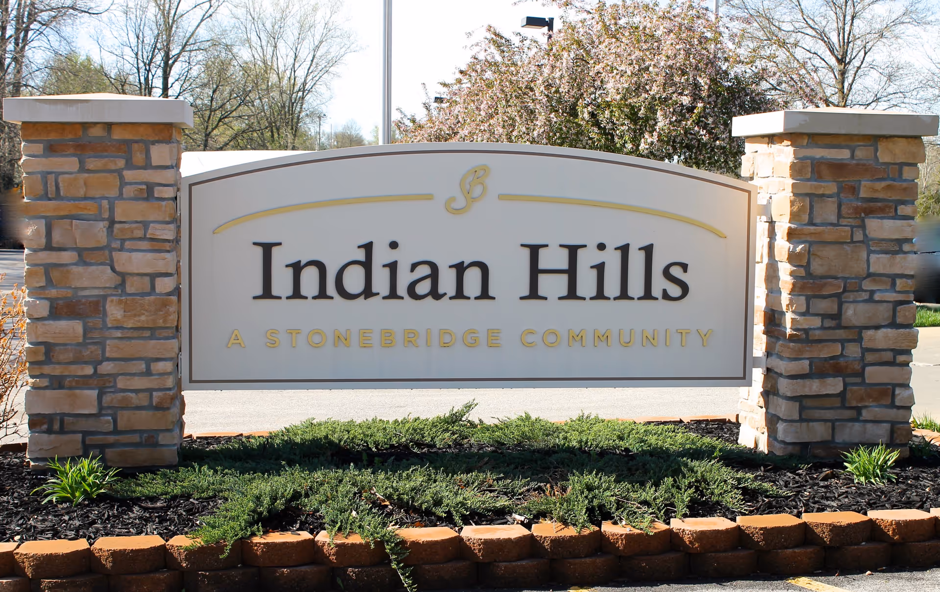 Outdoor stone sign for Indian Hills, a Stonebridge community, surrounded by landscaping with green shrubs and small plants, with trees and a clear sky in the background.