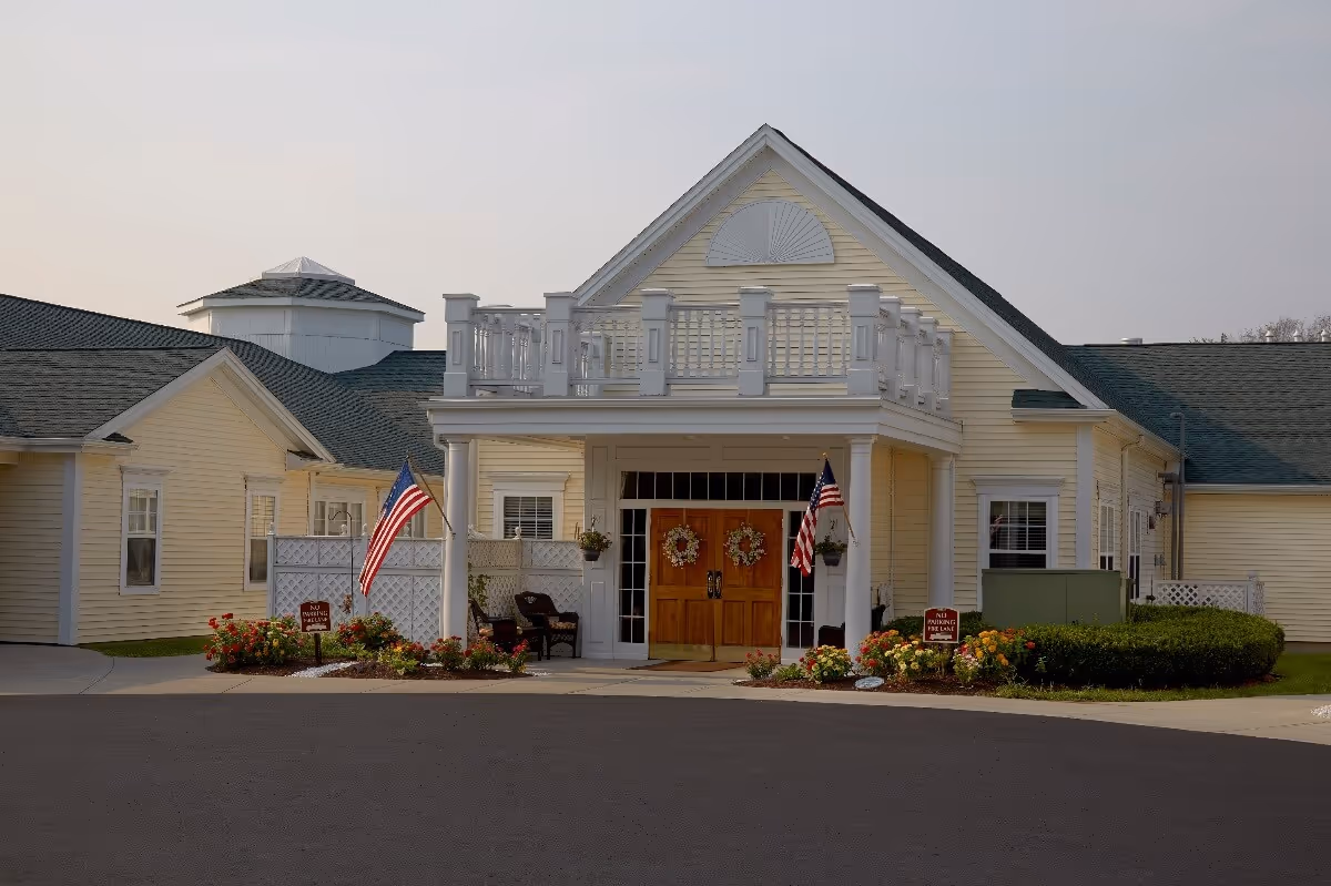 Front entrance of a pale yellow senior living facility with a covered porch, double wooden doors, American flags and landscaped flower beds.