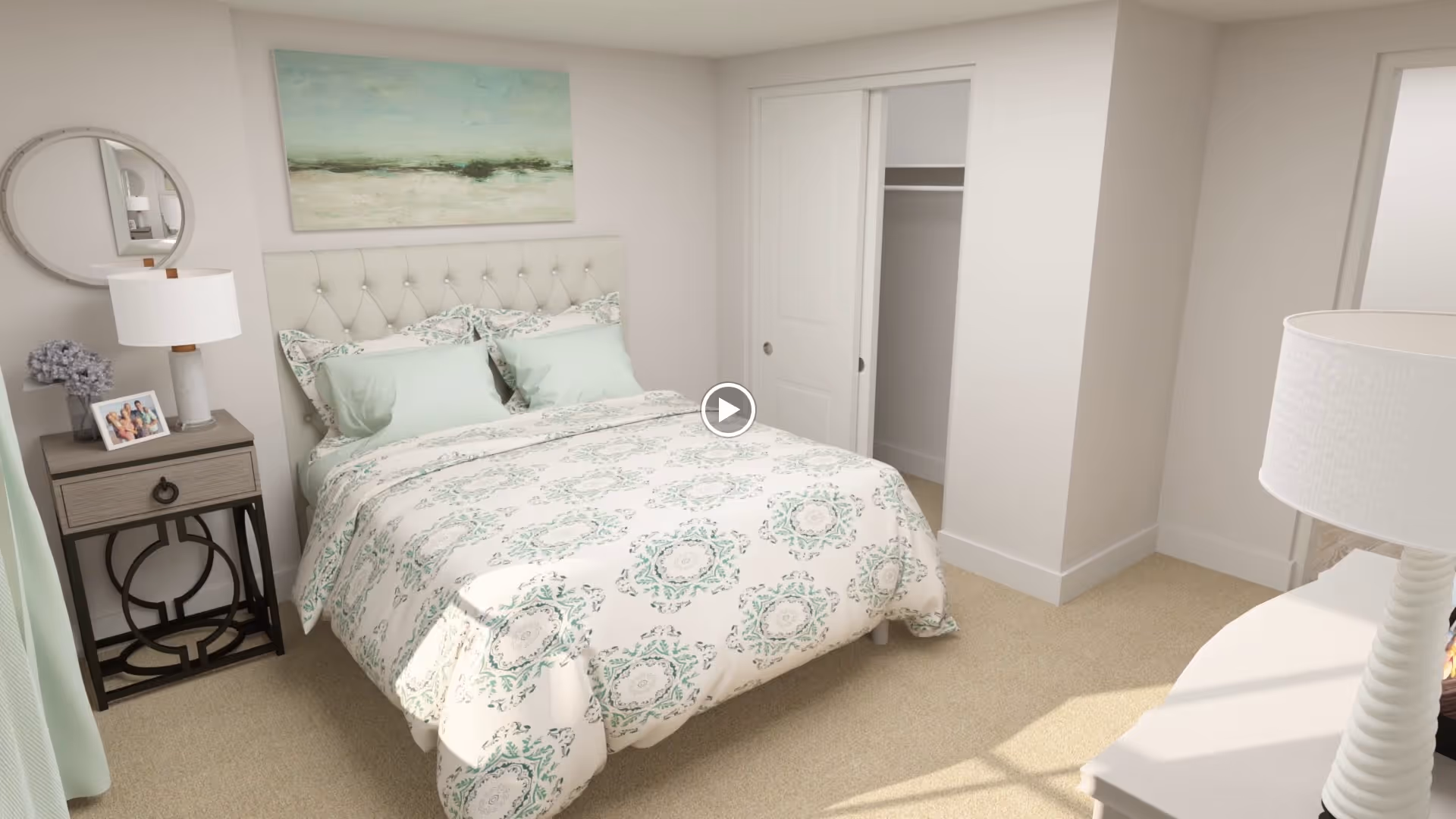 A cozy bedroom with a double bed featuring a tufted headboard and patterned bedding in soft green and white tones. There is a nightstand with a lamp, a small framed photo, and a vase with flowers. A round mirror hangs on the wall above the nightstand. The room has beige carpet flooring, a closet with sliding doors, and a white dresser with a lamp on it.