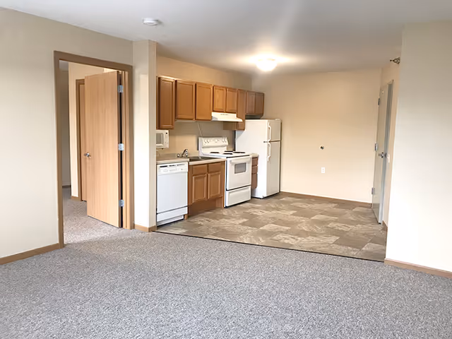 Interior view of a small kitchen area in a senior living facility with wooden cabinets, white appliances including a refrigerator, stove, microwave, and dishwasher. The kitchen floor is tiled, and the adjacent living area is carpeted. There is an open doorway leading to another room with an open wooden door.