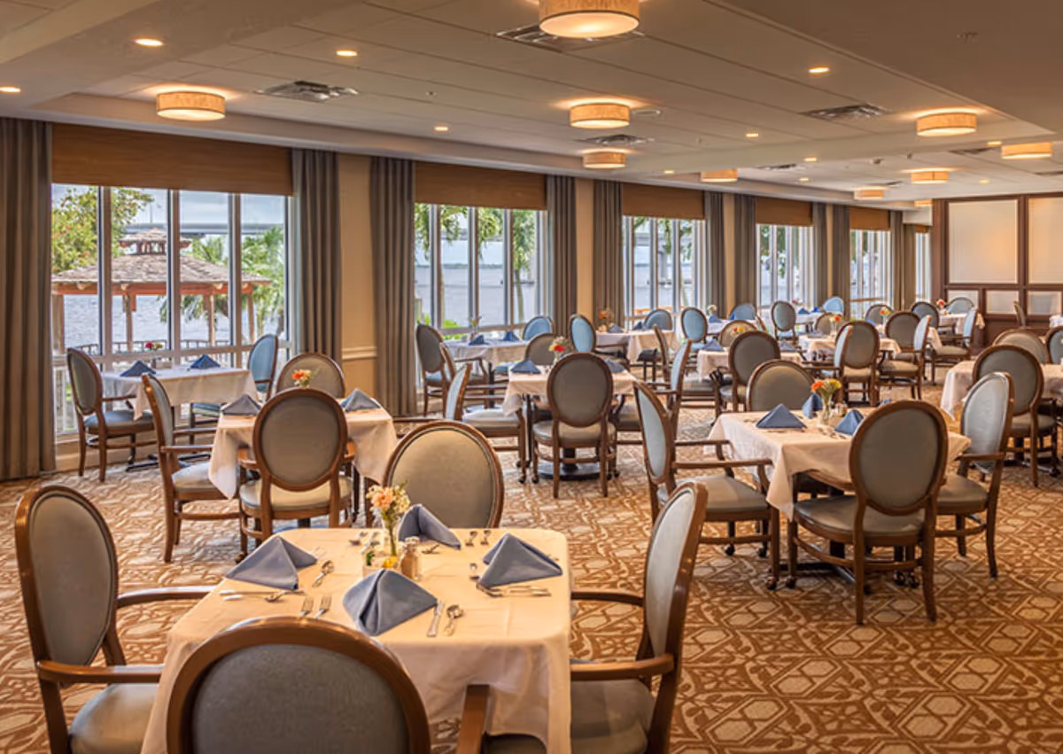 A spacious dining room with multiple tables covered in white tablecloths, each set with blue folded napkins, silverware, and small floral centerpieces. The room has large windows along one side offering a view of a waterfront and greenery outside. The chairs are wooden with cushioned seats and backs, and the ceiling features multiple round light fixtures.