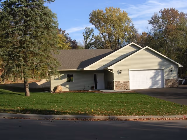 Single-story residential building with beige siding and a two-car garage, surrounded by green grass and trees with autumn foliage under a blue sky.
