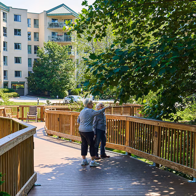 Two elderly women standing on a wooden walkway surrounded by lush green trees and foliage, with a multi-story residential building visible in the background on a sunny day.