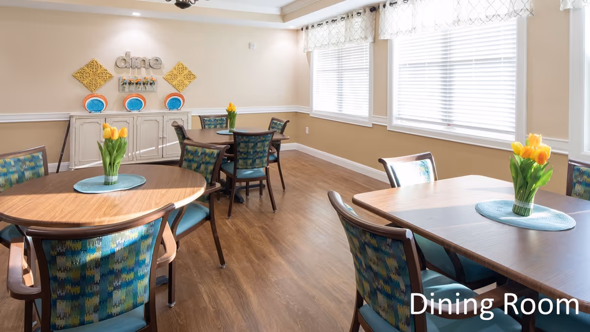 A bright dining room with wooden tables and chairs featuring blue and green patterned upholstery. Each table has a blue placemat and a vase with yellow tulips. The room has large windows with white blinds and light-colored walls decorated with wall art and plates. The word 'dine' is displayed on the wall above a white cabinet.