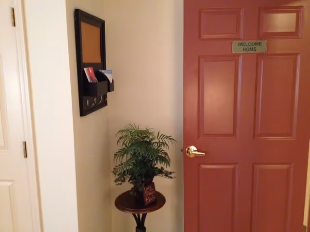 A red door with a green sign that says 'WELCOME HOME' on it. Next to the door is a small round table with a potted plant and a small sign. On the wall above the table is a black organizer with hooks and slots holding some papers.